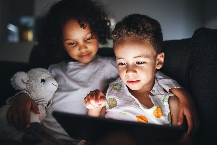 Two young children sitting on a couch in a dark room, faces lit by the glow of a tablet screen